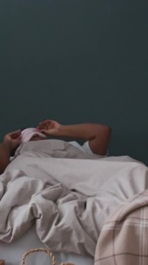 Dikey çekimde genç siyahi kadın uyanıyor, yatakta oturuyor, uyku maskesini çıkarıyor ve sabahları kendini zayıf hissediyor.