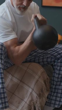 Yetişkin bir adamın evde yatakta oturup, sabah antrenmanı sırasında kettlebell bisepsi kıvrımları yaparken dikey pozu.
