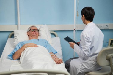 Doktor elinde tablet tutarken yaşlı hasta hastane yatağında dinleniyor. Tıbbi kayıtları inceliyor. Seruma bağlı hastayla parlak ayar ve tıbbi ekipmanlar görünür