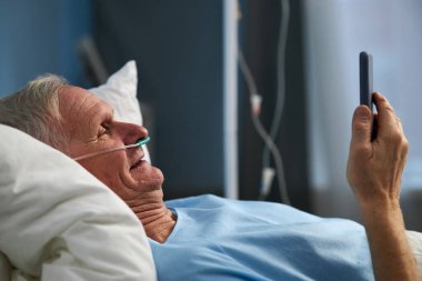 Yaşlı bir adam hastane yatağında uzanıyor. Bağlı kalmak ve eğlenmek için akıllı telefondan faydalanıyor. Arka planda tıbbi ekipman görülüyor. Hastane için ortam yaratılıyor.