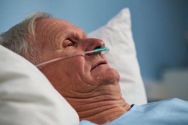 Hastane yatağında yatan kıdemli adam oksijen desteği için burun tüpü takıyor. Sağlık hizmetlerinde derin düşüncelere dalmış ve sakin görünüyor.