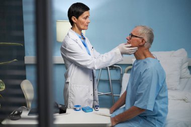 Beyaz önlüklü doktor hastane odasında yaşlı adamı muayene ediyor. Tıbbi malzemelerle dolu. Doğru teşhis ve hasta bakımını sağlama alıyor.