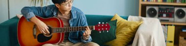 Asyalı bir adam kanepede oturmuş akustik gitar çalıyor rahat bir ortamda kitaplar ve arka planda bir teyp var.