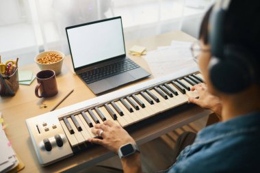 Çalışma istasyonunda dizüstü bilgisayarla elektronik klavye çalan bir adam ve çeşitli ofis malzemeleri etrafında müzikal bir çalışma ortamı yaratıyor.