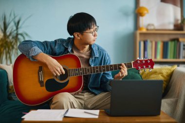 Asyalı müzisyen kanepede otururken gitar çalışıyor. Müzik notaları ve dizüstü bilgisayarla dikkatlice yan tarafa bakıyor.