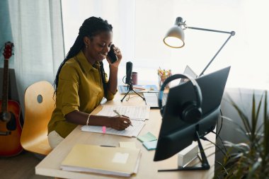 Afro-Amerikalı kadın telefonda konuşarak ve ofis ortamında masa başında notlar alarak birden fazla iş yapıyor. Arka planda müzik aletleri ve modern teknoloji görülüyor