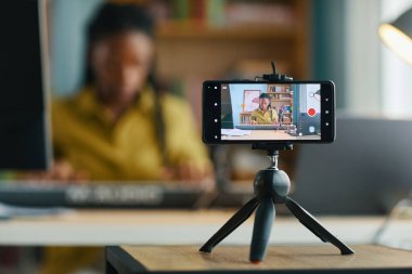 Ev ofis ortamında video içeriğini kaydetmek için akıllı telefonu tripoda kullanan bir kadın. Arkaplan bulanık, profesyonel video üretimi için aygıta odaklanmayı vurguluyor