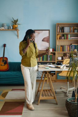 Ofiste telefon kullanan, ayakta duran bir kadın. Etrafı kitaplık, gitar ve masa ile çevrili. İşle ilgili bir konuşma yapıyor gibi görünüyor.