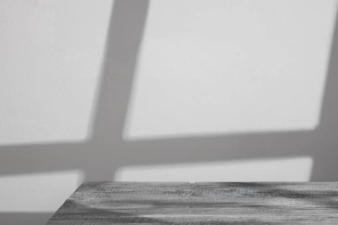 Beyaz bir duvarın karşısında beyaz ahşap bir masanın olduğu temiz ve minimalist bir fotoğraf. Pencerenin yumuşak gölgesi derinlik ve doku ekler, sakin ve sakin bir atmosfer yaratır.. 