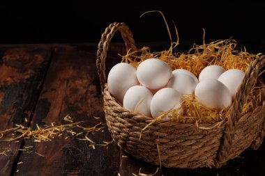 Beyaz yumurtalarla dolu örülmüş bir sepet, samanla dağılmış koyu ahşap bir yüzeyde oturur, kırsal, çiftlikten esinlenilmiş bir estetik yaratır..