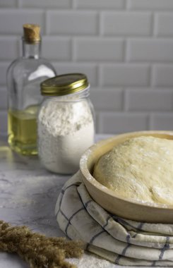 Ahşap bir kasede hamur mayası, cam bir kavanoz un ve hafif arka planda bir şişe yağ olan bir köy mutfağı sahnesi. Yumuşak, havadar tonlar pişirmek için rahat ve davetkar bir atmosfer yaratıyor.