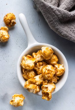 Kırsal bir daire, küçük beyaz bir kasede karamelli patlamış mısır ve açık gri beton arka planda etrafa saçılmış parçalar. Sıcak altın renkler, rahat bir his için dokulu yüzeyle tezat oluşturur.