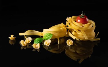 Çiğ tagliatelle makarnası, kiraz domatesli, fesleğen yapraklı ve parlak siyah bir yüzeyde küçük makarna parçaları. İtalyan mutfağı, ambalajı veya menü tasarımı için ideal minimalist besin bileşimi. 