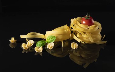 Çiğ tagliatelle makarnası, kiraz domatesli, fesleğen yapraklı ve parlak siyah bir yüzeyde küçük makarna parçaları. İtalyan mutfağı, ambalajı veya menü tasarımı için ideal minimalist besin bileşimi. 