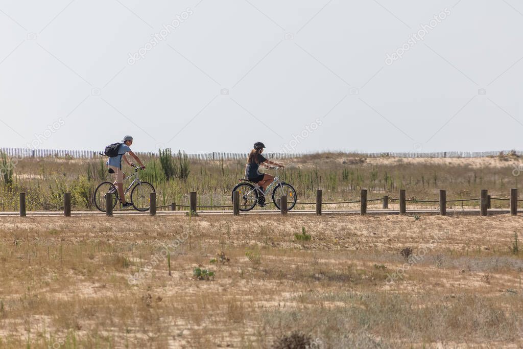 Figueira da Foz Portugal 08 09 2022: Couple riding bikes, on the Claridade  beach dunes on bike path / pedestrian walkways, dunes and vegetation  typical of the Portuguese coast as background 2023
