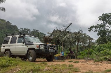 Sumbe Angola - 02 23 2020: Geleneksel meyve satan bir kadının dış görünüşü, palmiye dalları ve ahşap gövdelerden yapılmış çadırında, yan tarafta 4X4 araç, arkada tropikal orman, Sumbe, Angola