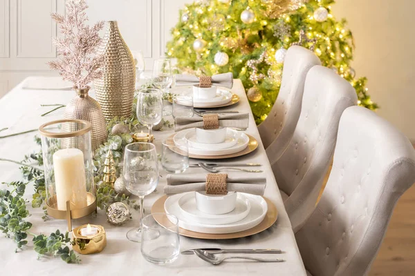 Table set for Christmas dinner, dinnerware set on gold charger plates in white and gold colors with candles, inside a Scandinavian house interior