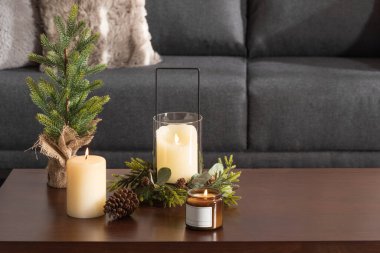 Noel süslemesi, mumlar ve köknar ağacı dallarıyla ahşap bir masaya yakın, rahat bir oturma odasında, arka planda gri bir kanepe