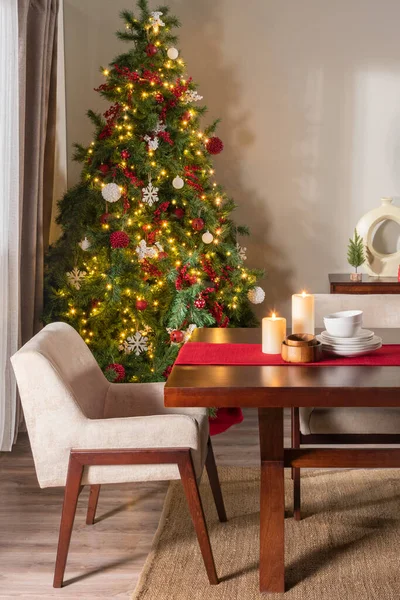 Decorated Norway artificial Christmas tree in the Nordic, warm dining room with candles and tableware set on a wooden table with a fabric chair. Christmas celebration at home