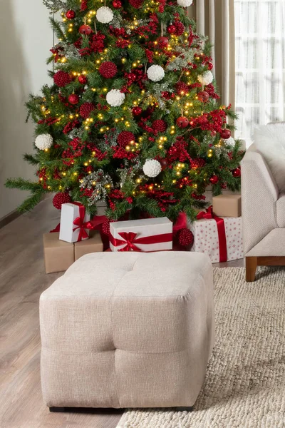 Living Room in an Apartment Decorated for the Holidays with a Christmas Tree Adorned with Red Ornaments and Festive Lights, Gifts, and a Rectangular Pouf, Evoking a Warm Holiday Atmosphere