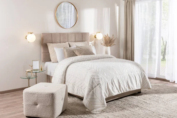 Modern Scandinavian-Style Bedroom with Neutral Tones, Featuring a Double Bed with White Cover, Rectangular Pouf, and Mirror, Bathed in Neutral Daylight from a Window