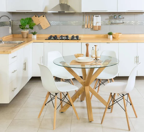 Modern Scandinavian Kitchen, Featuring a Dining Area with a Circular Glass Dining Table Set and Plastic Chairs with Wood Finish, Kitchen Cabinet with Bamboo Countertop and Cooking Accessories.