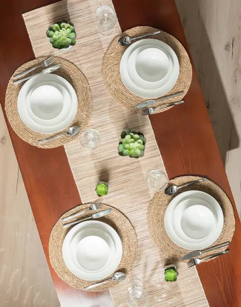 Elegant Wooden Table Setting with Ceramic White Plates on Round Placemats and Table Runner, Accented with Wine Glasses and Small Decorative Potted Plants, Perfect for a Party or Dinner - Top View.