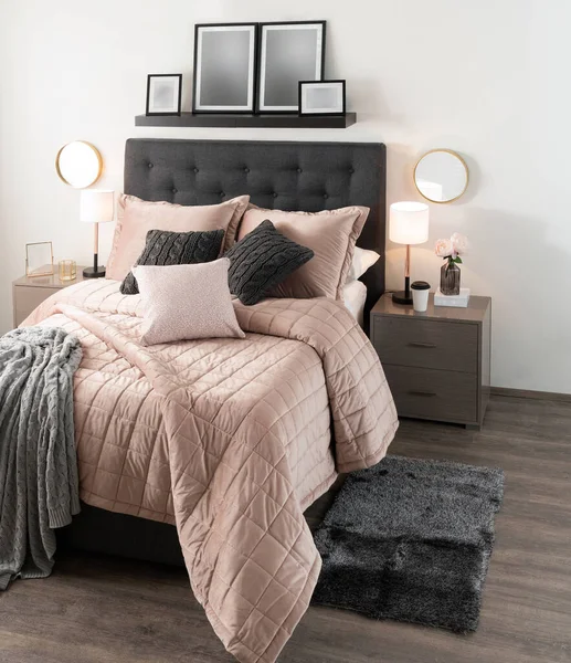Bedroom Interior Featuring a Comfortable Bed with Pink Luxury Diamond Pattern Comforter, White Cozy Pillows, and Grey Blanket, a Picture Ledge Shelf on a White Wall, Beige Side Tables, Natural Light.