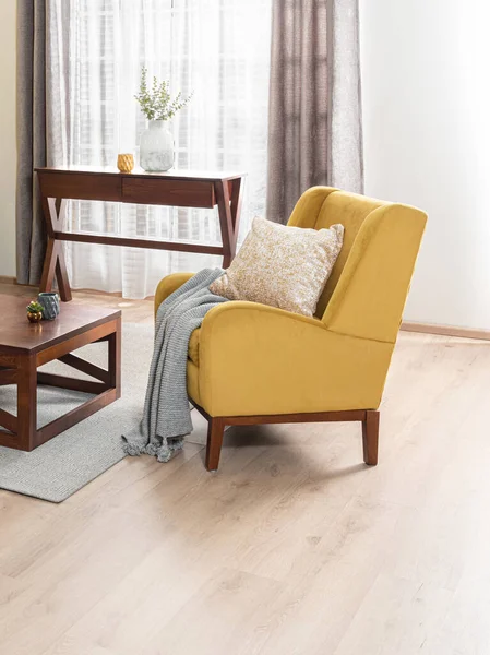 Yellow Mid-Century Modern Fabric Lounge Armchair Adorned with a Beige Decorative Pillow in a Modern Living Room Illuminated by Natural Light from the Window and Wooden Coffee Table, Wooden Flooring.