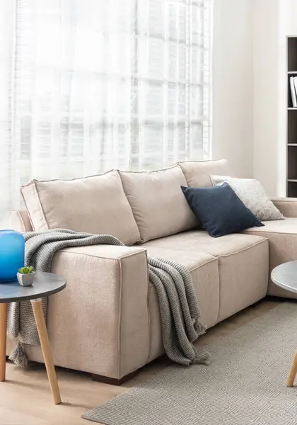 Modern Minimal Living Room with Beige Fabric Corner Sofa, Carpet, and Coffee Table, Features Textured Decorative Cushions and a Throw Blanket for the Couch, set Illuminated by a Window.