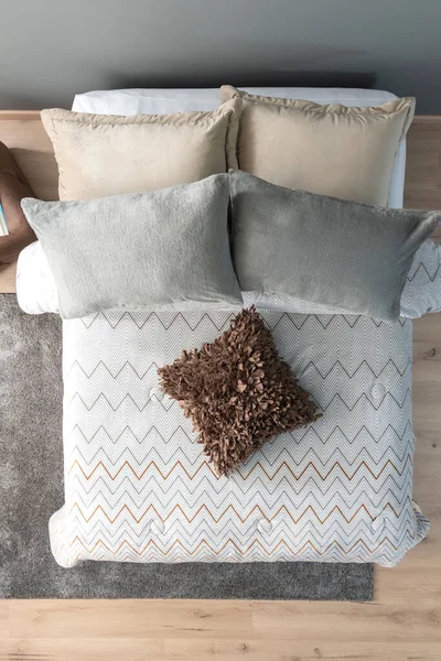 Contemporary Bedroom, Fluffy Gray Pillows atop Crisp White Bed Linen, Beige Cushion, A Chevron-Patterned Throw with a Chunky Brown Textured Pillow, Gray Wall, Wooden Floor and Area Rug, Top View.