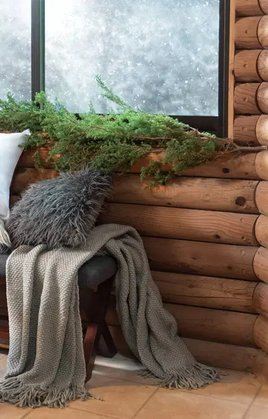Winter Cabin Interior: Fluffy Grey Cushion and Intricately Woven Blanket Artfully Draped Over a Decorative Fabric Bench, Wooden Wall with Green Pine Branches, a Frosted Window, Christmas Ambience.