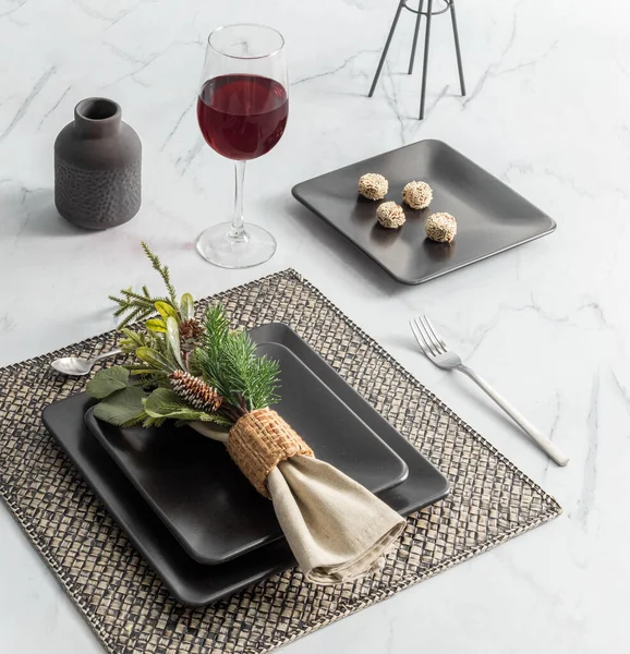 Modern minimalist dining setup on a marble table featuring sleek matte-black square plates, delicate truffles, an elegant glass, a rustic napkin decoration, and a placemat, festive Christmas dinner.