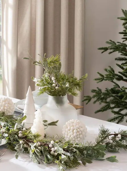Nordic Christmas decor in a serene setting, glass vase holding green branches, white ceramic Christmas tree and berry ornaments on a table, featuring shimmering garlands, soft beige curtains, sunlight