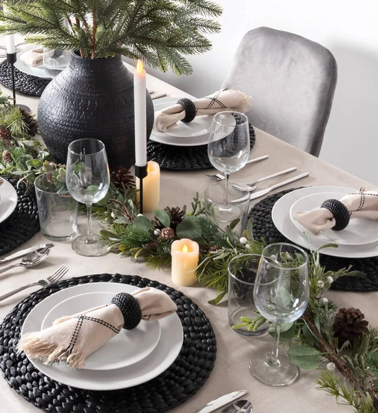 Festive New Year\'s Eve dinner table adorned with a pine garland centerpiece, decorative woven black placemats, elegant white dishes with fringed napkins and candles. white wall and grey dining chair.