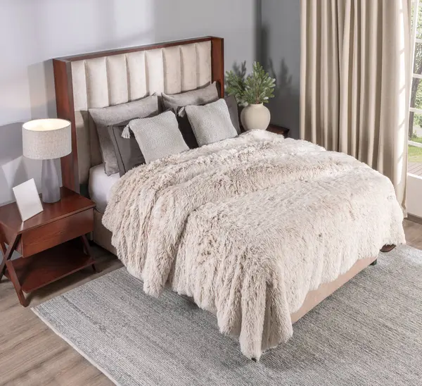 A Nordic bedroom featuring a plush, textured beige fur blanket on bed, wooden headboard with cream upholstery, pillows, a wooden nightstand with a picture frame, and curtains beside a sunlit window.