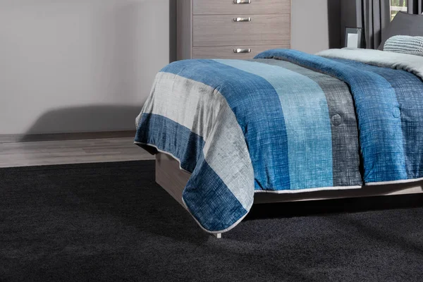 Youthful bedroom setting: close-up of a modern bed with a blue and white striped quilt, grey wooden base, deep charcoal carpet, and a sleek wooden dresser with metallic handles. Nordic style.