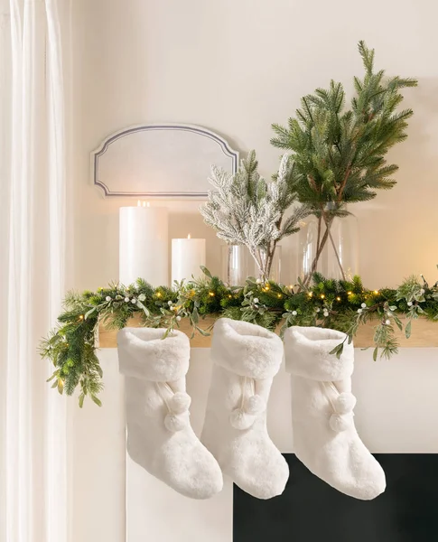 Christmas Hearth, White Plush Christmas Stockings Hung on a Fireplace Mantel Adorned with Pine Garland, Glittering Lights, Pillar Candles, Complemented by a White Wall, Natural Light, Festive Decor.