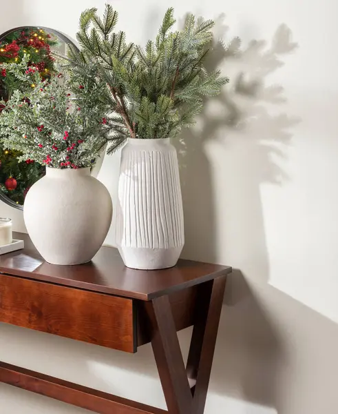 Festive Foyer Display, Mahogany Console Table with a Round White Ceramic Vase and a Textured White Pottery Vase Holding Sprigs of Green Fir and Red Berry Branches, Reflecting in a Round Mirror.
