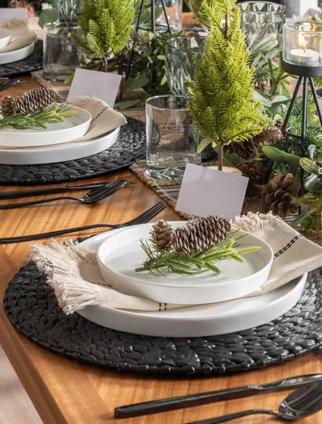 Rustic Nordic Holiday Table: Black Cutlery on Textured Placemats, White Porcelain Dishes with Pine Cone Accents, Miniature Evergreen Centerpieces, and Burlap Napkins, Set Upon a Solid Wood Table.