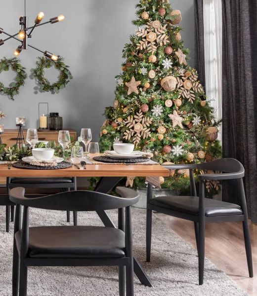 Scandinavian Christmas Dining, Contemporary Black Chairs, Natural Oak Table, Set with Woven Placemats, Pristine Porcelain Bowls, Crystal Stemware, Lush Fir Tree Adorned with Stars, Festive Atmosphere.