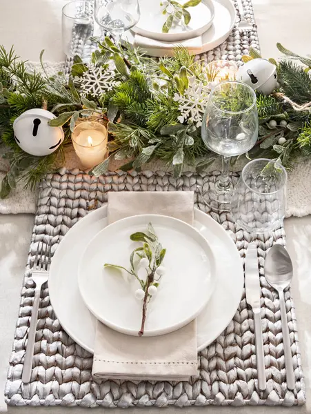 Scandinavian Table Setting Aesthetic: Simple White Plates on Textured Woven Placemats, Paired with Sleek Silverware, Amidst a Rustic Greenery Garland with Snowflake Accents and Soft Candlelight.