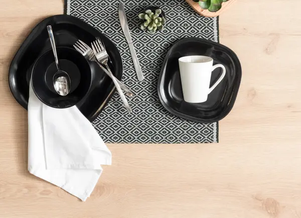Contemporary Dining Elegance, Top View of a Chic Table Setting with Black Dinnerware, White Linen Napkin, Polished Silverware, on a Geometric Patterned Placemat with a Textured Wooden Backdrop.