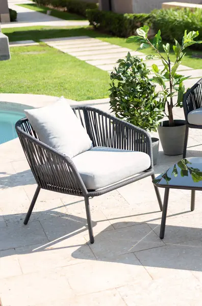 Sun-Drenched Modern Patio Elegance with a Black Wire Armchair, Grey Cushions, Beside a Contemporary Side Table and Verdant Potted Plants, Overlooking the Calm Waters of a Private Home\'s Pool.