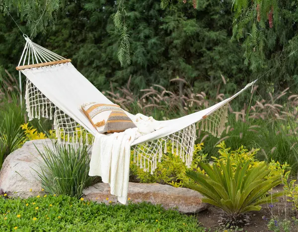 Tranquil Garden Oasis Featuring a White Cotton Hammock with Fringe Detail, Adorned with a Striped Cushion and Knit Throw, Nestled Among Lush Greenery and Vibrant Yellow Flowers, with a Majestic Fern.