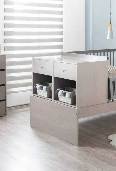 Contemporary Nursery Room Featuring a Modern Convertible Crib and a Matching Wood Grain Laminate Dresser with Built-in Cubbies and Woven Baskets, Grey Wooden Flooring and Stylish Striped Window Shades