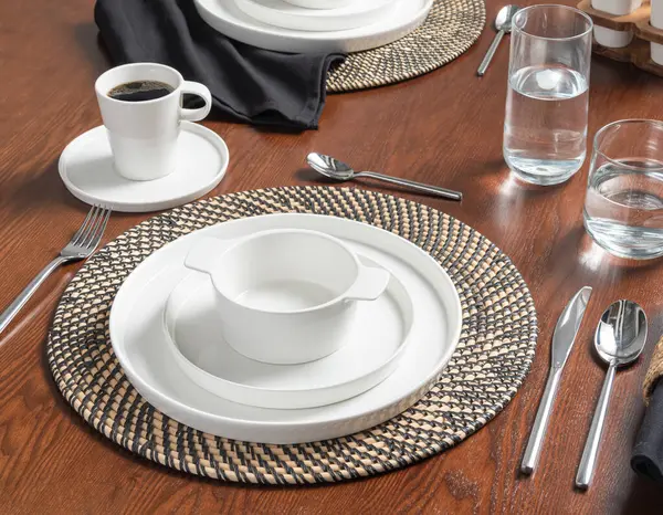 stock image Brunch Table Setting Featuring Elegant White Porcelain Dishware on Woven Rattan Chargers, Stainless Steel Flatware and Clear Glass Drinkware, Freshly Brewed Cup of Coffee on a Rich Wooden Dining Table