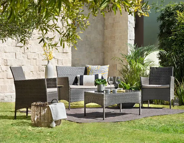 stock image Sunlit garden dining space, a rattan weave furniture set, two armchairs and a sofa with neutral cushions, a coffee table against a textured stone wall backdrop, refreshing morning meal lush greenery.