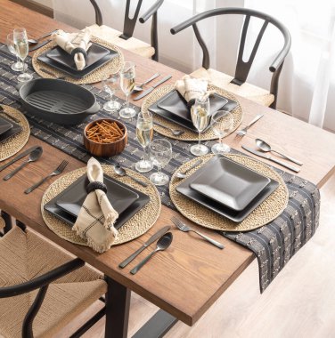 Top view of a dining table set for a meal, featuring modern black dinnerware on rustic woven placemats, complemented by elegant glassware, metallic utensils, and a stylish wooden table, well-lit room. clipart