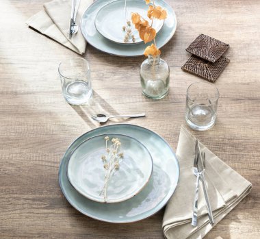 Top view of an elegantly set dining table, light blue ceramic plates, simple glassware and woven coasters, set on a rustic wooden table textile napkins, floral decor, Art Deco-inspired dining ambiance clipart